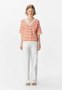 Striped V-neck top in beige and orange, paired with white wide-legged pants. Fabric appears soft with a relaxed fit. Sandals complete the look.