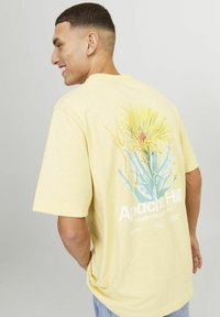 T-shirt in cotone giallo con una grafica floreale sul retro, in colori verde e giallo, e il testo "Apache Hill" in bianco.