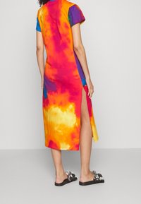 Šaty s tie-dye vzorem v živých oranžových, růžových, modrých a fialových barvách, s krátkými rukávy a bočním rozparkem, vyrobené z měkké tkaniny.