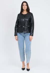 Giubbotto di pelle nera con collo rotondo, chiusura frontale, due tasche a toppa e dettagli metallici. Indossato sopra jeans azzurri chiari e scarpe nere.