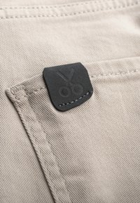 Beige stof met een gestructureerd oppervlak, voorzien van een zwarte leren label in de vorm van scharen, bevestigd aan een zak met zichtbare stiksels.