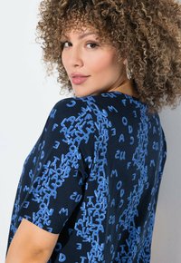 Černé tričko s výrazným opakujícím se modrým typografickým vzorem. Krátké rukávy a volný střih. Hladká bavlněná textura.