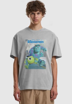 Młody mężczyzna noszący jasnoszary T-shirt z kolorowymi postaciami z filmu „Potwory i spółka” - Mike i Sulley biegnący oraz napisem „Monsters” z przodu.