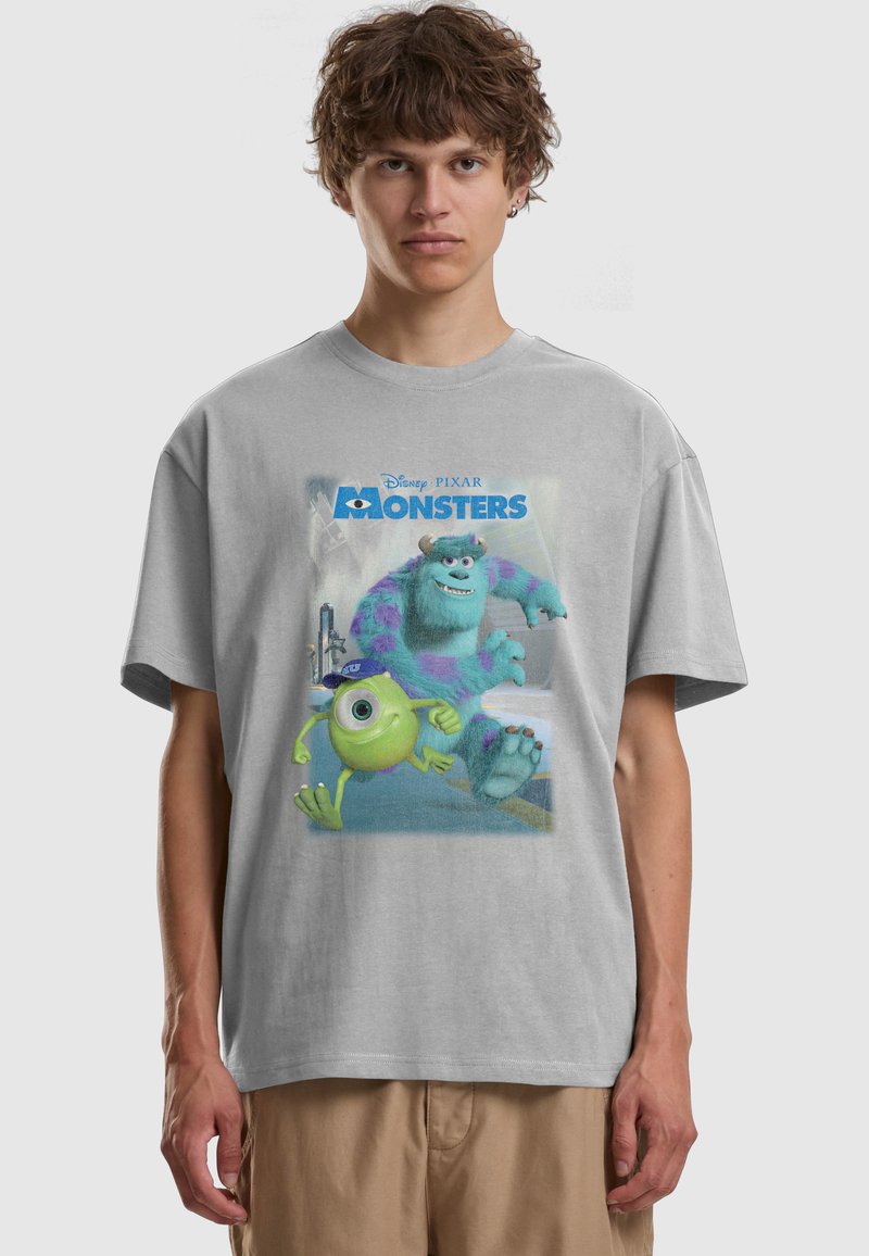 Jeune homme portant un t-shirt gris clair avec les personnages colorés de Monsters, Inc. Mike et Sulley courant, et le texte "Monsters" sur le devant.