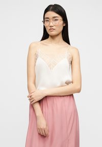 Νεαρή γυναίκα με μακριά σκουρόχρωμα μαλλιά, που φορά διαφανή γυαλιά, λευκό αμάνικο τοπ με δαντελένιες λεπτομέρειες και ροζ φούστα, στέκεται με σταυρωμένα τα χέρια.