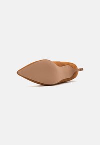 Zapato de tacón alto de ante en marrón claro con punta afilada. Cuenta con una suela beige lisa y una pequeña correa trasera para facilitar su uso.