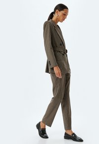 Braun kariertes Blazer mit Taillengürtel, kombiniert mit passenden Hosen. Glänzende schwarze Loafer runden das Outfit ab. Glatte Textur, maßgeschneiderte Passform.