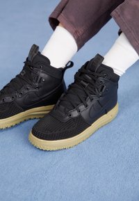 Person som bär svarta högkaresneakers med beige sulor, vita strumpor och mörkröda byxor på ett blått mattgolv.