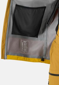 Gelbe Jacke, geöffnet, um die innere Netztasche und die Stoffmerkmale zu zeigen, mit einem Etikett, das wasserdicht, atmungsaktiv, winddicht, recycelte Materialien, verklebte Nähte und PFC-frei hervorhebt.