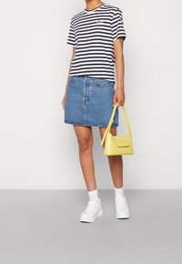 Person som bär en svart och vit randig t-shirt, blå denimkjol, vita sneakers och håller en liten gul handväska.