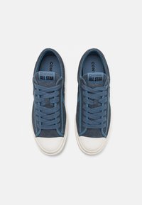 Zapatillas de gamuza azul con suela de goma blanca, con cordones planos, detalles de costuras y una etiqueta de marca en la lengüeta.