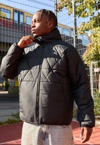 Quilted sort jakke med lynlås foran og høj krave. Den har en glat overflade og to sidelommer, båret over lyseblå jeans.