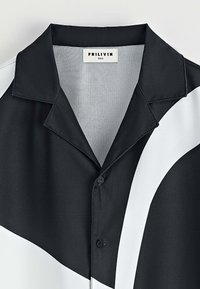 Chemise noire et blanche à manches courtes, avec un col, texture satinée douce, fermeture boutonnée et motif diagonal distinct. Étiquette de marque visible.