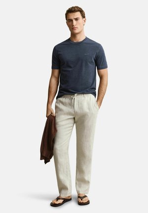 Mann trägt dunkelblaues kurzärmeliges T-Shirt, helle beige Kordelzughose, braune Flip-Flops, hält braunen Pullover in der linken Hand.