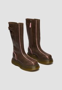 Kõrged pruunid nahast saapad roosade vertikaaltriipudega, külgedel tõmblukud ja paksud läbipaistvad tallad kollaste õmblustega, seisavad helehalli tausta peal.