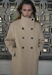 Beżowy wełniany płaszcz z wysokim kołnierzem, dwurzędowym zapięciem na czarne guziki oraz bocznymi kieszeniami, na tle ściany z wzorem.