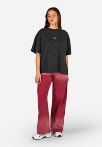 Camiseta preta oversized com um pequeno logótipo gráfico centrado no peito, combinada com calças largas em degradê rosa e ténis brancos.