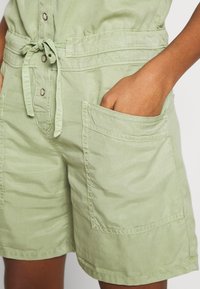 Main dans la poche d'une combinaison verte claire à boutons avec ceinture à nouer et grandes poches avant sur une personne à la peau bronzée.