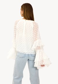 Blouse blanche avec manches à volants, en tissu transparent avec motifs floraux embossés. Associée à un jean bleu clair, vue de dos.