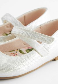 Paire de chaussures Mary Jane argentées étincelantes avec finition texturée, doublure intérieure fleurie et brides ajustables à scratch, présentée sur fond blanc.