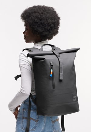 Frau mit Afro-Frisur, die ein weißes Langarmshirt und blaue Jeans trägt und einen großen schwarzen Rolltop-Rucksack auf dem Rücken hat.
