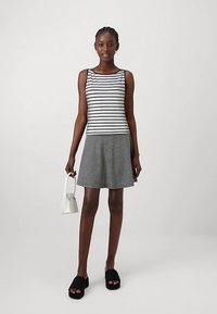 Débardeur rayé noir et blanc associé à une jupe à carreaux. Le modèle tient un petit sac à main blanc et porte des sandales noires à enfiler.