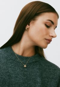 Kuld sõlm kõrvarõngad ja sobiv ripats kaelakee. Kõrvarõngastel on tekstureeritud, põimunud disain. Modell kannab tumerohelist kootud kampsunit.