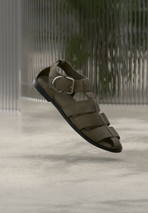 Καφέ δερμάτινο σανδάλι με πολλαπλούς ιμάντες και ασημένια αγκράφα, απεικονίζεται σε θολό φόντο με υφή γυαλιού πάνω σε λεία επιφάνεια.