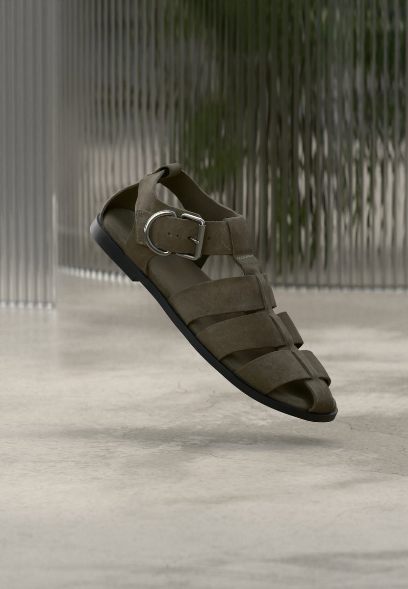 Καφέ δερμάτινο σανδάλι με πολλαπλούς ιμάντες και ασημένια αγκράφα, απεικονίζεται σε θολό φόντο με υφή γυαλιού πάνω σε λεία επιφάνεια.