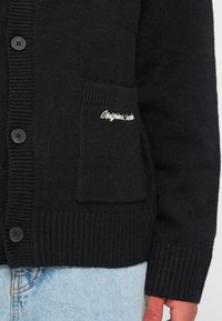 Čierny cardigan s rebrovanou textúrou, s gombíkmi a prednou vreckom vyšitým nápisom "Originals". Vyrobený z hrubého, mäkkého materiálu.