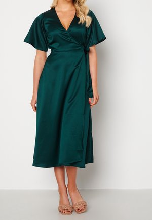 Femme portant une robe portefeuille en satin vert foncé avec des manches courtes évasées et des talons ouverts beiges tressés, debout contre un mur blanc.