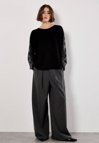 Donna che indossa un top a maniche lunghe nero con maniche decorate e pantaloni grigi a righe larghe, in piedi contro un muro bianco.