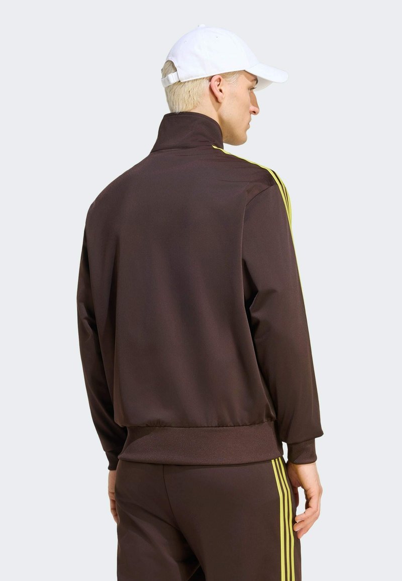 Uomo che indossa una tuta marrone con strisce gialle e un cappellino bianco, in piedi e di profilo su uno sfondo uniforme.