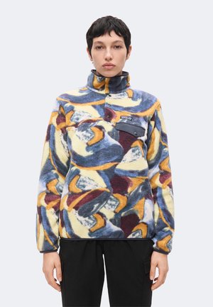 Junge Person mit kurzen dunklen Haaren, die eine mehrfarbig abstrakt gemusterte Fleecejacke mit hohem Kragen und schwarzen Hosen trägt.