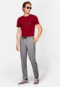 Czerwona koszulka z grafiką roweru, szare spodnie w kratkę ze sznurkiem w talii oraz szare trampki. Okulary przeciwsłoneczne dopełniają całość.