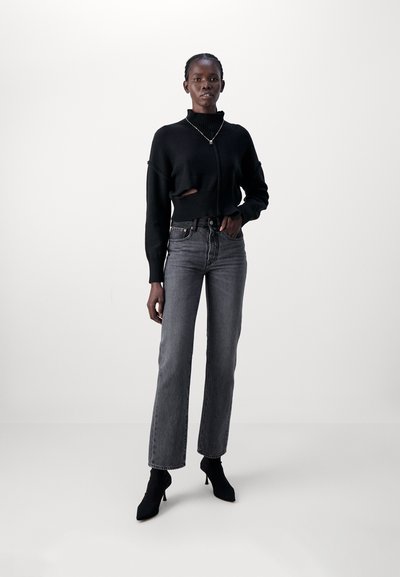 Black cropped sweater with a high neckline and cut-out detail, paired with high-waisted gray jeans and pointed black heeled boots.
