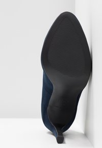 Semelle d'une chaussure à talon bleu avec un bout pointu et une semelle extérieure en caoutchouc noir texturé, positionnée contre un coin de mur et de sol blancs.