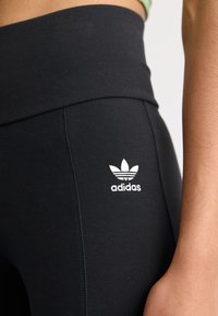 Melni legingi, izgatavoti no elastīga materiāla, ar augstu jostasvietu un baltu Adidas logo tuvu gurnam, ar gludu, matētu tekstūru.