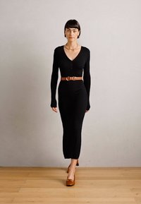 Black ribbed knit dress with long sleeves, V-neck, and button detail. Accented with a tan belt. Worn with brown shoes. Hardwood floor background.