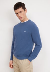 Blauwe gebreide trui met een gestructureerd patroon, ronde halslijn en geribde manchetten. Bevat een klein logo op de borst. G dragen met beige broek.