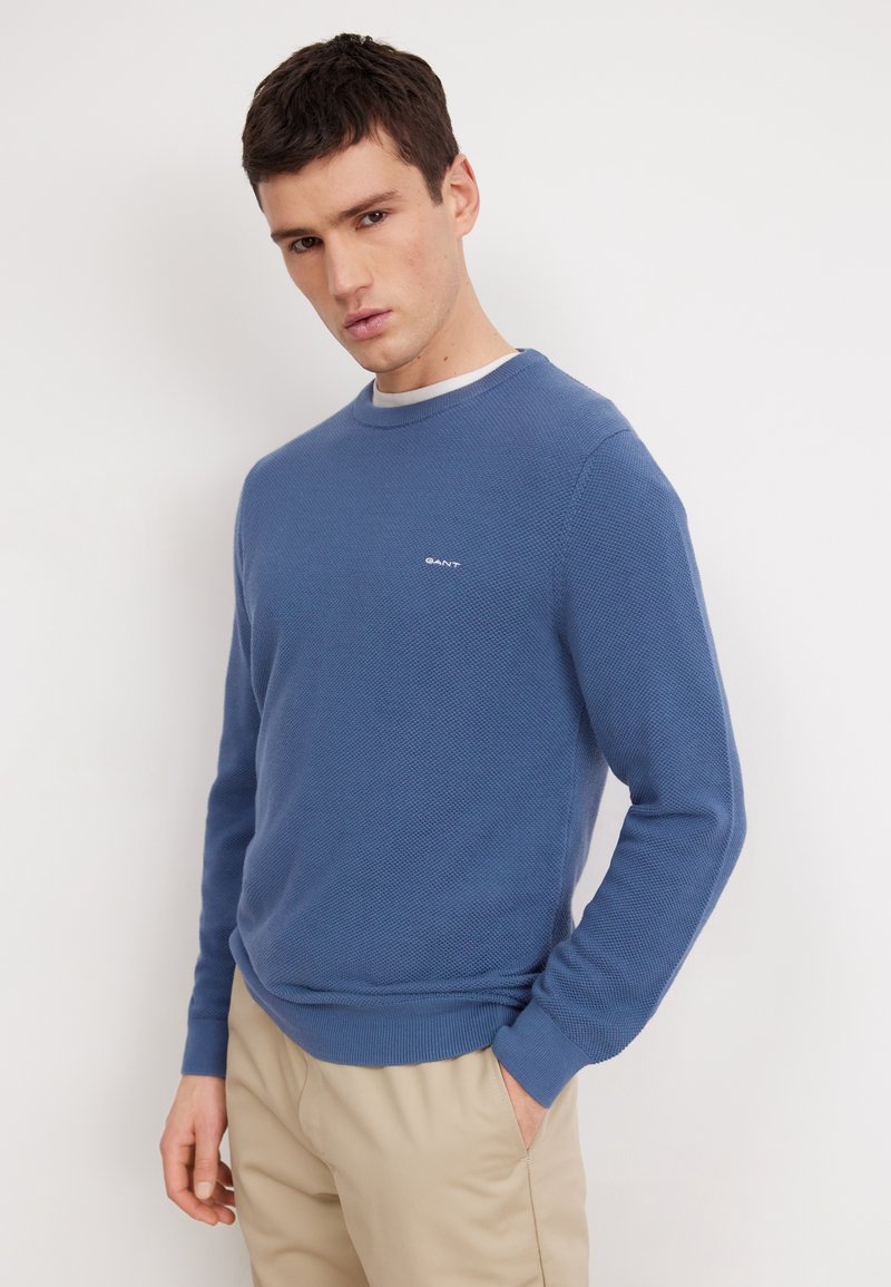 Blauwe gebreide trui met een gestructureerd patroon, ronde halslijn en geribde manchetten. Bevat een klein logo op de borst. G dragen met beige broek.
