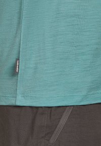 Una camiseta de manga corta en color teal hecha de un tejido ligero y texturizado, con una pequeña etiqueta que dice "Icebreaker" cerca del dobladillo, combinada con pantalones oscuros.