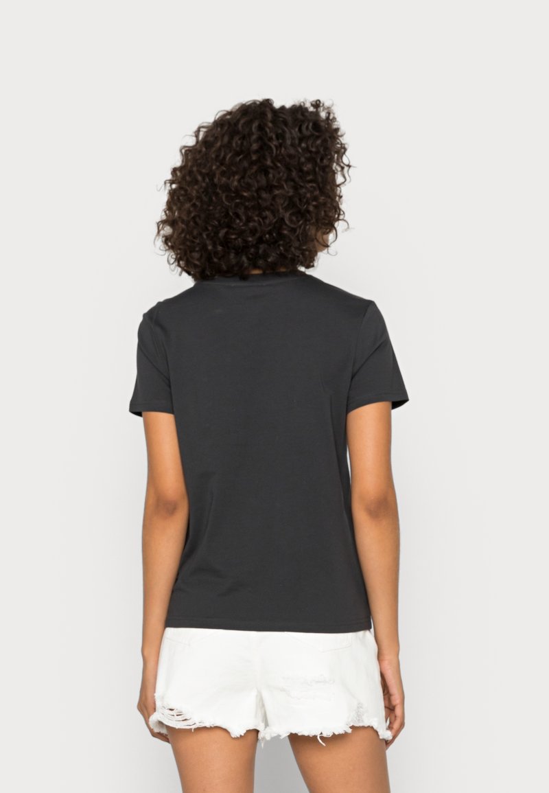 Femme aux cheveux bouclés portant un t-shirt noir à manches courtes et un short blanc déchiré, debout de dos devant un fond uni.