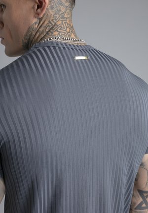 Man met nektattoos en zilveren ketting, gekleed in een grijs geribd shirt met een klein metalen label aan de bovenkant van de rug.