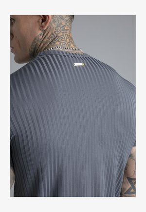 Nyakán tetoválásokkal és ezüst lánccal rendelkező férfi, szürke bordázott ingben, amelyen egy kis fémtábla látható a hát felső részén.