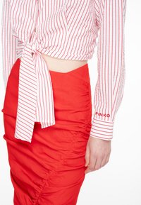 Camicia bianca con righe verticali rosse, annodata in vita, abbinata a una gonna rossa arricciata. Le maniche presentano un dettaglio ricamato "PINKO".