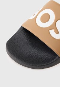 Brūna gumijas slaidu sandale ar teksturētu melnu zolīti un platu siksnu, uz kuras virsmas ir izteiksmīgi balti burti.