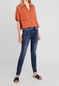 Camisa corta de color óxido con dos bolsillos en el pecho, combinada con jeans oscuros que presentan una franja roja a los lados. Sandalias planas negras completan el look.