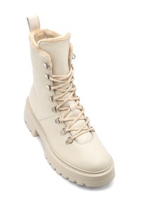 Kremowy skórzany botek za kostkę z sztucznym futerkiem, metalowymi oczkami i kremowymi sznurówkami; masywna podeszwa dla stabilności.