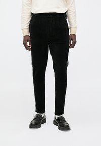 Pantalon en velours côtelé noir à coupe ajustée, présentant des rainures verticales, des bandes latérales et une fermeture bouton, associé à des mocassins noirs.